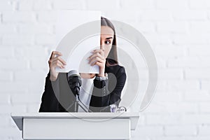 young lecturer suffering from fear of public speaking hiding face with paper sheet while standing on podium tribune.