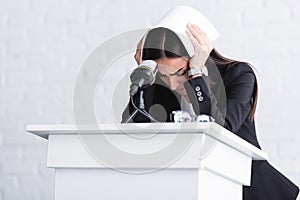 young lecturer, suffering from fear of public speaking, standing on podium tribune and covering head with paper.