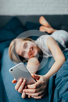 Young light brown hair woman in bed at home. Happy  girl smiling while texting message on mobile telephone.  Teenager lying in bed