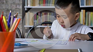 Young little asian boy painting on paper and sitting behind table, kid doing homework, book shelves background