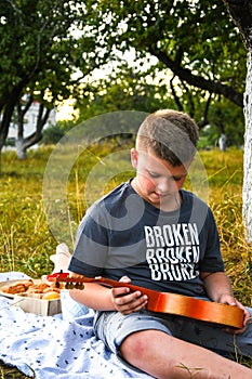 Young little boy guitarist outdoor. Boy on city park summer meadow enjoying day playing guitar