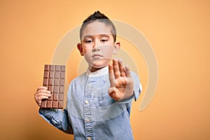 Young little boy kid eating sweet chocolate bar for dessert over isolated yellow background with open hand doing stop sign with