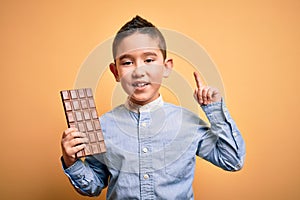 Young little boy kid eating sweet chocolate bar for dessert over isolated yellow background surprised with an idea or question