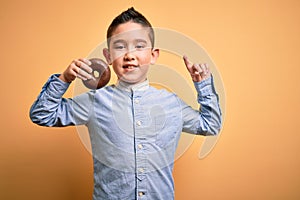 Young little boy kid eating unhealthy chocolate doughnut over isolated yellow background surprised with an idea or question