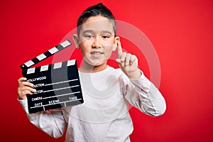 Young little boy kid filming video holding cinema director clapboard over isolated red background surprised with an idea or