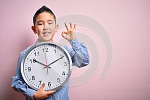 Young little boy kid holding big minute clock over isolated pink background doing ok sign with fingers, excellent symbol