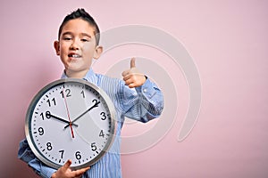 Young little boy kid holding big minute clock over isolated pink background happy with big smile doing ok sign, thumb up with