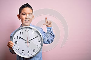 Young little boy kid holding big minute clock over isolated pink background surprised with an idea or question pointing finger