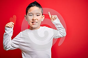 Young little boy kid holding heart paper shape over isolated red background surprised with an idea or question pointing finger