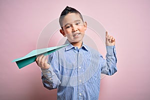 Young little boy kid holding paper plane over isolated pink background surprised with an idea or question pointing finger with