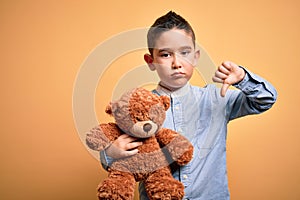 Young little boy kid hugging teddy bear stuffed animal over yellow background with angry face, negative sign showing dislike with