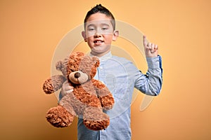 Young little boy kid hugging teddy bear stuffed animal over yellow background surprised with an idea or question pointing finger