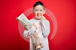 Young little boy kid looking at turist city destination map over red isolated background annoyed and frustrated shouting with