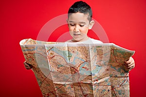 Young little boy kid looking at turist city destination map over red isolated background with a happy face standing and smiling