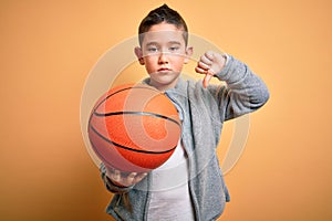 Young little boy kid playing with basketball game ball over isolated yellow background with angry face, negative sign showing