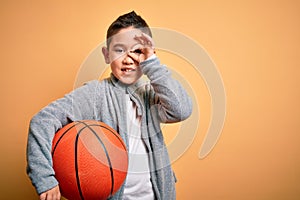 Young little boy kid playing with basketball game ball over isolated yellow background with happy face smiling doing ok sign with