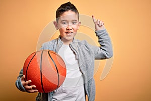 Young little boy kid playing with basketball game ball over isolated yellow background surprised with an idea or question pointing