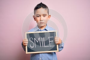 Young little boy kid showing blackboard with smile word as happy message over pink background with a confident expression on smart