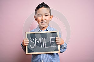 Young little boy kid showing blackboard with smile word as happy message over pink background with a happy face standing and