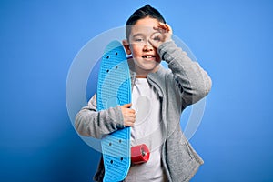 Young little boy kid skateboarder holding modern skateboard over blue isolated background with happy face smiling doing ok sign