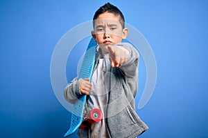 Young little boy kid skateboarder holding modern skateboard over blue isolated background pointing with finger to the camera and