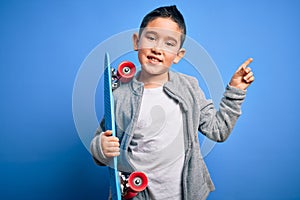 Young little boy kid skateboarder holding modern skateboard over blue isolated background very happy pointing with hand and finger