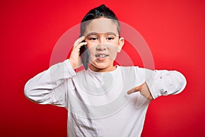 Young little boy kid talking on smartphone mobile over red isolated background with surprise face pointing finger to himself