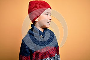 Young little boy kid wearing wool cap and winter sweater over yellow isolated background looking away to side with smile on face,