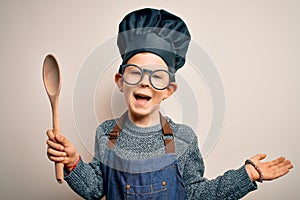 Young little caucasian cook kid wearing chef uniform and hat cooking using wooden spoon very happy and excited, winner expression