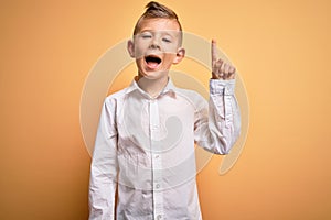 Young little caucasian kid with blue eyes wearing elegant white shirt over yellow background pointing finger up with successful