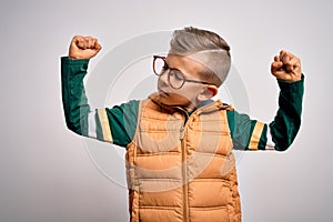 Young little caucasian kid with blue eyes wearing winter coat and smart glasses showing arms muscles smiling proud