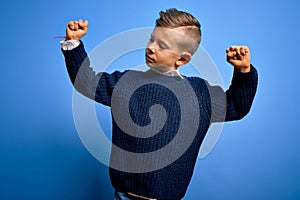 Young little caucasian kid with blue eyes wearing winter sweater over blue background showing arms muscles smiling proud