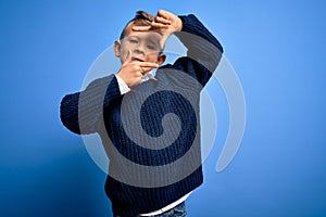 Young little caucasian kid with blue eyes wearing winter sweater over blue background smiling making frame with hands and fingers