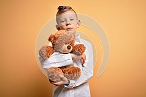 Young little caucasian kid hugging teddy bear stuffed animal over yellow background with a confident expression on smart face