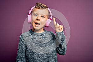 Young little caucasian kid wearing headphones listening to music over purple background pointing finger up with successful idea