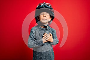 Young little caucasian kid wearing vintage biker motorcycle helmet and googles over red background smiling with hands on chest