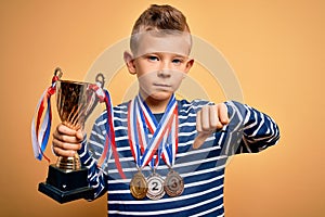 Young little caucasian kid wearing winner medals and victory award trophy over yellow background with angry face, negative sign