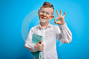 Young little caucasian student kid wearing smart glasses and holding a book from school doing ok sign with fingers, excellent