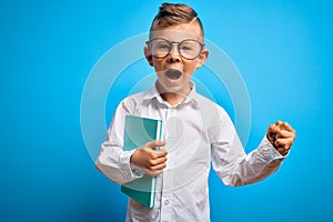 Young little caucasian student kid wearing smart glasses and holding a book from school screaming proud and celebrating victory