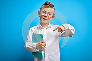 Young little caucasian student kid wearing smart glasses and holding a book from school with surprise face pointing finger to