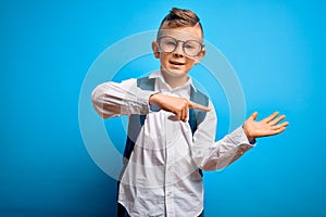 Young little caucasian student kid wearing smart glasses and school bag over blue background amazed and smiling to the camera