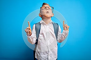 Young little caucasian student kid wearing smart glasses and school bag over blue background amazed and surprised looking up and