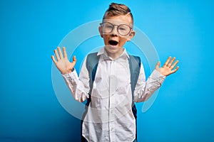 Young little caucasian student kid wearing smart glasses and school bag over blue background celebrating crazy and amazed for