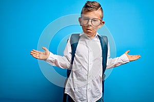 Young little caucasian student kid wearing smart glasses and school bag over blue background clueless and confused expression with