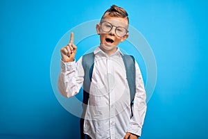 Young little caucasian student kid wearing smart glasses and school bag over blue background pointing finger up with successful