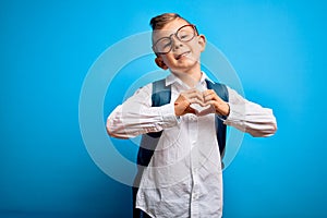 Young little caucasian student kid wearing smart glasses and school bag over blue background smiling in love showing heart symbol