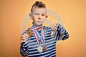 Young little caucasian winner kid wearing award competition medals over yellow background with angry face, negative sign showing