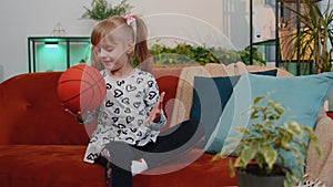 Young little children girl basketball fan holding ball looking at camera, training dribbling at home