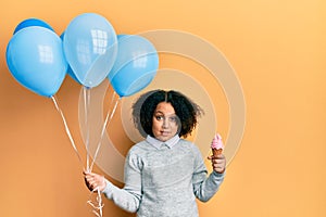 Young little girl with afro hair holding ice cream and blue balloons clueless and confused expression