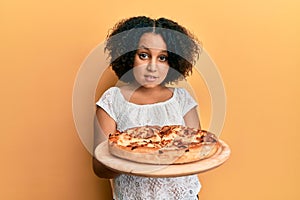 Young little girl with afro hair holding italian pizza clueless and confused expression